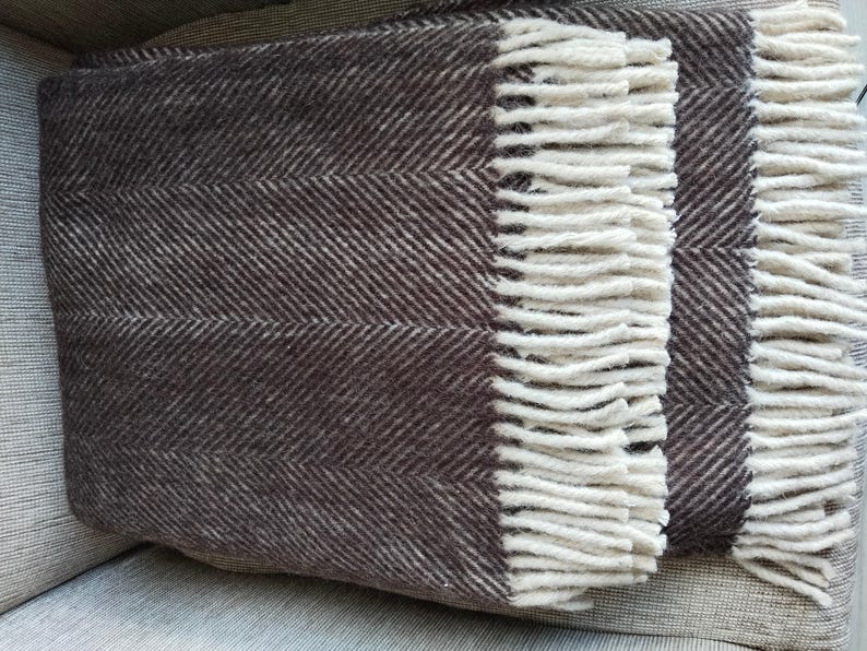 May include: A folded brown and white herringbone pattern throw blanket with cream-colored fringe. The blanket is made of a soft, textured material and is folded neatly. The fringe adds a decorative touch to the blanket's edge.