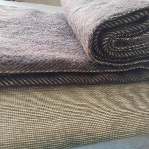May include: A folded brown and beige wool blanket with a herringbone pattern. The blanket has a fringed edge and is resting on a textured, light brown surface. The blanket is folded in a layered fashion.