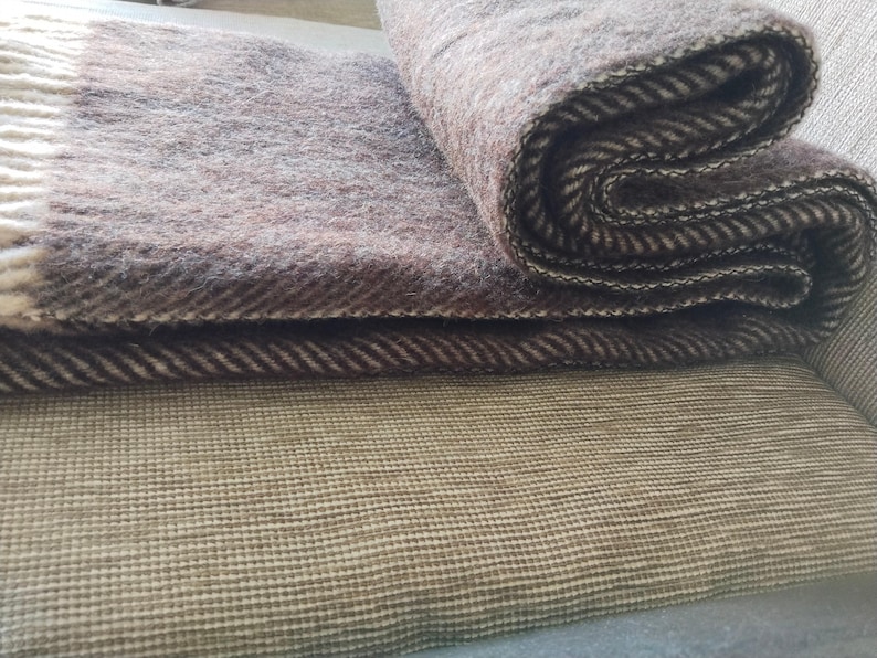 May include: A folded brown and beige wool blanket with a herringbone pattern. The blanket has a fringed edge and is resting on a textured, light brown surface. The blanket is folded in a layered fashion.
