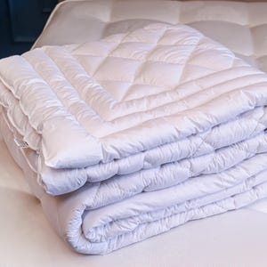 May include: A stack of folded white quilted blankets on a white mattress. The blankets have a diamond pattern and are neatly folded. The bedding is clean and appears soft, suggesting comfort and quality.