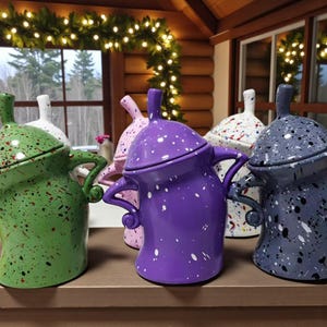 May include: A collection of colorful ceramic teapots with lids and handles. The teapots are in various colors, including green, purple, pink, white, and gray, each with a speckled paint design. The teapots are displayed on a wooden surface.