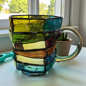 以下が含まれることがあります： 透明なハンドルが付いた、カラフルなステンドグラスのマグカップ。マグカップは、黄色、緑、青、オレンジ、赤のガラス片を黒で縁取ったパッチワークデザインです。マグカップは白い表面に置かれています。