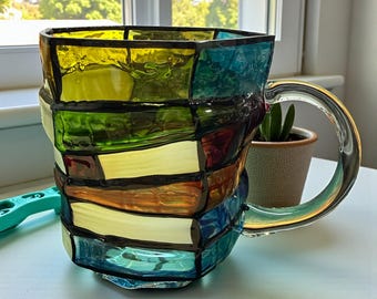 May include: A colorful stained glass mug with a clear handle. The mug features a patchwork design of yellow, green, blue, orange, and red glass pieces, outlined in black. The mug is sitting on a white surface.
