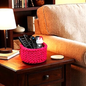 May include: A vibrant hot pink, chunky knit circular basket made of thick yarn. The basket is filled with several black electronic remote controls and a small white device, serving as a functional storage container for living room accessories.