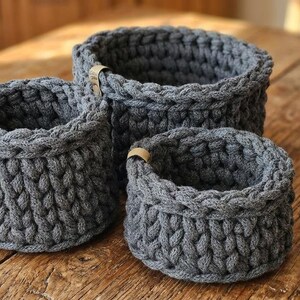 May include: Three gray, hand-crocheted baskets of varying sizes are arranged on a wooden surface. Each basket has a rounded shape and a textured, woven appearance. Small leather tags are attached to the rims. The baskets are a neutral home decor item.