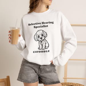May include: A white long-sleeve sweatshirt featuring a black graphic of a Cavoodle puppy. The text above the puppy reads Selective Hearing Specialist and the text below reads CAVOODLE. The sweatshirt is paired with gray denim shorts.