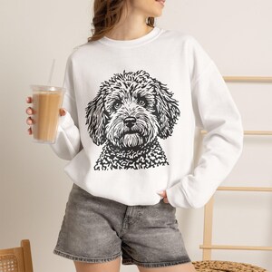 May include: A white long-sleeve sweatshirt featuring a black graphic illustration of a curly-haired dog on the front. The sweatshirt has a crew neckline and ribbed cuffs, paired with gray denim shorts. The design depicts a detailed dog face.