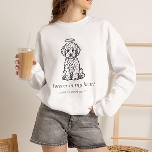 May include: A white long-sleeve sweatshirt featuring a black line drawing of a curly-haired dog with a halo above its head. Below the illustration, the text reads, "Forever in my heart" and "until we meet again" in a simple black font.