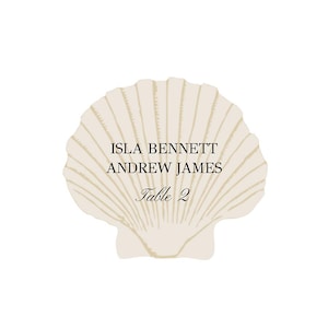 May include: A seashell-shaped place card with the names "ISLA BENNETT" and "ANDREW JAMES" printed in black. Below the names is the text "Table 2" in a script font. The seashell is a light beige color with detailed lines.