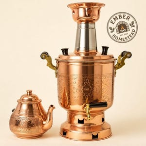 May include: A polished copper samovar with an ornate engraved floral pattern, featuring a silver-toned chimney, brass handles, and a black spigot handle. Beside it sits a matching copper teapot with similar floral engravings and a lid.
