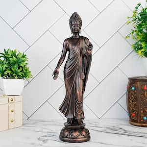 May include: A bronze-effect Buddha statue in a standing pose, with one hand raised. The statue has detailed robes and hair, and stands on a lotus flower base. The background is a white tiled wall. The statue is approximately 30 cm tall.