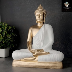 Puede incluir: Estatua de Buda dorada y blanca en posición de meditación sentado. La estatua presenta una cabeza y brazos dorados, con una túnica blanca sobre los hombros. La base es dorada y la estatua está sobre un fondo oscuro.