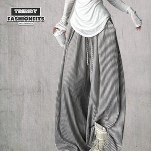 Puede incluir: Pantalones anchos a rayas verticales grises y blancas con cintura con cordón, combinados con una camiseta blanca de manga larga. La imagen incluye el texto "TRENDY FASHIONFITS" y "UNISEX FASHION FITS".
