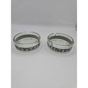 May include: Two clear glass ashtrays with a circular design. Each ashtray has a dark green band around the rim and a decorative border with a repeating pattern of figures. The ashtrays are on a white surface.