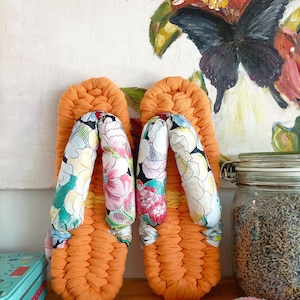Peut inclure: Une paire de sandales tressées orange dotées de brides épaisses et rembourrées, ornées d'un motif floral coloré dans des tons rose, vert et blanc. Les sandales présentent une base tressée texturée et une silhouette traditionnelle de type zori.