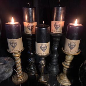 May include: Five black candles with brown paper labels that say "devil brand" and a cartoon image of a devil. The candles are on black and brown candle holders.