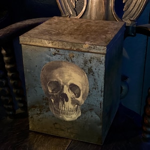 May include: A vintage metal box with a skull design. The box is painted in a distressed blue and brown color scheme. The skull is a black and white image.