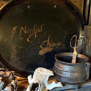 May include: A vintage still life composition. A large, circular, black metal tray with gold lettering reading "Needful Things" is displayed. A small cast iron cauldron, scissors, and a skull are also visible.