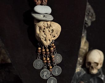 May include: A handcrafted necklace featuring a brown leather cord, small wooden beads, and a pendant made of stacked stones and a porous beige stone. The pendant is adorned with dangling silver-toned coins. The necklace is displayed on a black surface.