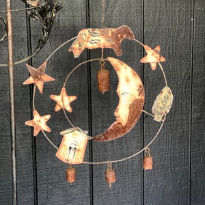 May include: A metal wind chime with a crescent moon, stars, a house, and a cow. The chime has bells hanging from it and is rusty brown in color.