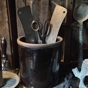 May include: A collection of vintage kitchen tools, including cleavers, knives, and scissors, are stored in a dark brown ceramic crock. A large wooden spoon and other antique items are visible in the background, creating a rustic aesthetic.