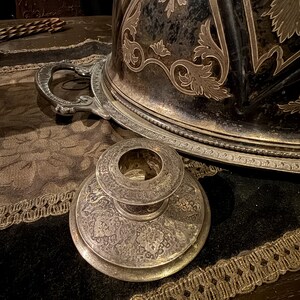 May include: An antique silver-plated serving dish with ornate floral and scrollwork designs. The lid is domed and features a handle. A matching candlestick holder is in the foreground. The items are on a dark surface with gold trim.