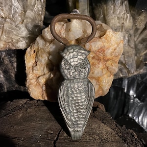 May include: A silver owl bottle opener with a rusty metal ring. The opener is sitting on a piece of wood.