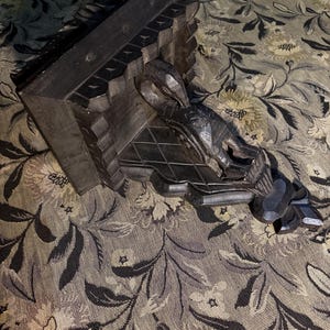 May include: A dark brown, carved wooden shelf bracket. The bracket features a decorative design with geometric and stylized elements. The shelf is L-shaped and appears to be made of solid wood. The bracket is designed to be mounted on a wall.