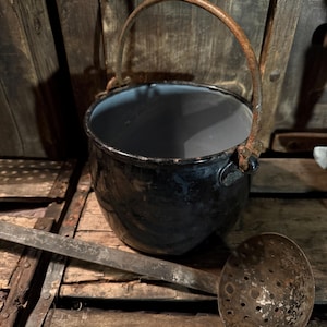 May include: A black enamel pot with a rusty metal handle and a matching ladle. The pot has a blue interior and sits on a wooden surface. The ladle has a perforated bowl and a long handle. The scene evokes a rustic, vintage aesthetic.