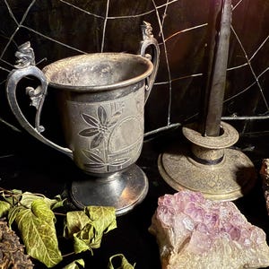 May include: An antique silver trophy cup with floral engravings and ornate handles. A black candle burns in a vintage candlestick, next to a cluster of amethyst crystals and dried ivy. The scene is set against a dark backdrop.