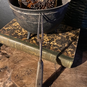 May include: A vintage, rustic, metal, two-pronged fork with a wooden handle. The fork is resting on top of a closed, antique book with a brown leather cover. The book is sitting on a wooden surface.