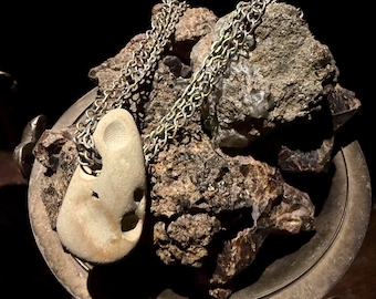 May include: A stone pendant necklace with a silver chain rests on a bed of rough, dark brown rocks inside a silver-colored bowl. The pendant is a light beige color with a unique, organic shape and a hole.