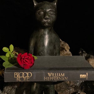 May include: A black book titled "Blood Rose" by William Heffernan, with a red rose on top of it, and a black cat statue in the background.