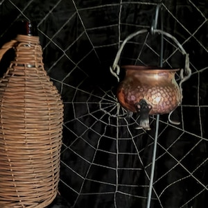 May include: A brown wicker-covered bottle and a small copper cauldron with a handle, suspended from a metal stand. The background features a spiderweb design against a dark backdrop, creating a Halloween theme.