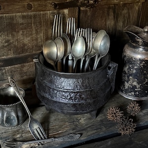 May include: A collection of vintage silverware, including forks and spoons, is displayed in a black cast iron cauldron. The cauldron sits on a weathered wooden surface, alongside a small silver dish and a decorative pitcher. The scene evokes a rustic, antique aesthetic.