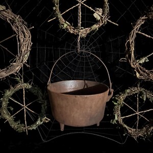 May include: A brown cast iron cauldron with three short legs and a curved metal handle, positioned in front of a black wire spiderweb structure decorated with five circular wreaths made of brown twigs and green leaves.