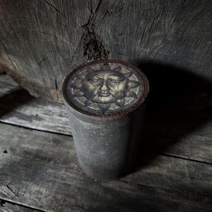 May include: A cylindrical metal container with a detailed sun face design on the lid. The container has a weathered, aged appearance, with a dark, muted color palette. The background is a wooden surface.