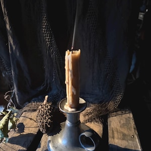May include: A lit, beige candle with melting wax sits in a silver candlestick holder. Smoke rises from the candle against a dark backdrop of draped fabric and a wooden surface. A pinecone and foliage are also visible.