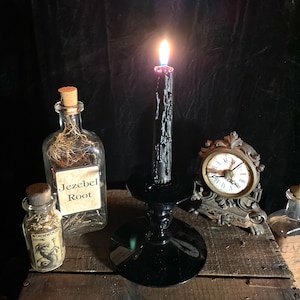 May include: A black candle burning on a black candlestick, with a glass bottle labeled "Jezebel Root" and a smaller bottle labeled "Wormwood" on a wooden crate.