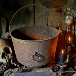 May include: A large, rusty metal cauldron with a handle and ring detail, set against a rustic wooden backdrop. A skull, a decorative doll, a saw, and lit candles complete the spooky, vintage aesthetic.