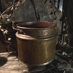 May include: A hammered copper cauldron with a curved handle. The pot has a wide, cylindrical body and a slightly flared rim. The metal has a warm, aged patina. The background includes a wooden surface and foliage.
