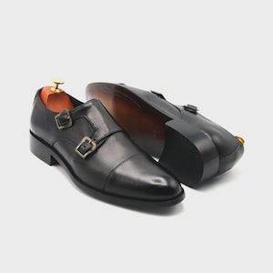 Puede incluir: Zapatos de cuero negro con doble hebilla y hebillas doradas. Los zapatos tienen un dise&ntilde;o de puntera y se muestran con hormas de madera en el interior. Las suelas son de color marr&oacute;n oscuro.