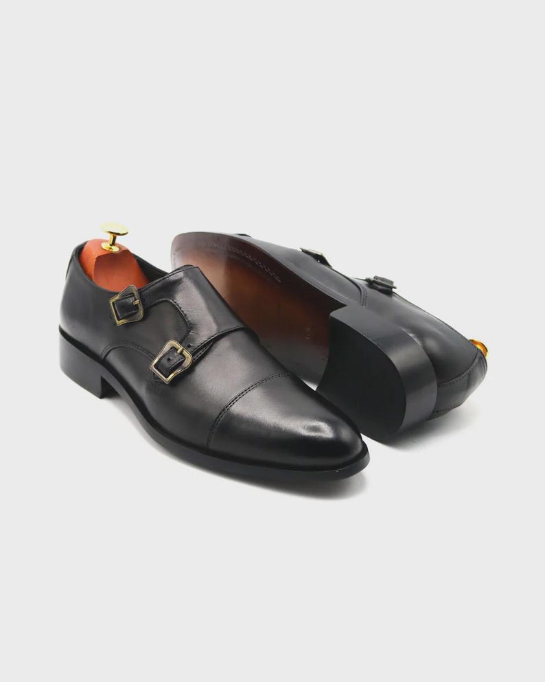 Puede incluir: Zapatos de cuero negro con doble hebilla y hebillas doradas. Los zapatos tienen un dise&ntilde;o de puntera y se muestran con hormas de madera en el interior. Las suelas son de color marr&oacute;n oscuro.