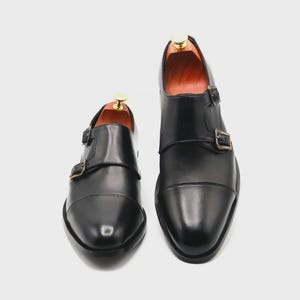 Puede incluir: Un par de zapatos negros de cuero con doble hebilla. Los zapatos tienen un dise&ntilde;o de puntera y dos hebillas ajustables. Hormas de madera en el interior. Se muestran sobre un fondo blanco. Estilo cl&aacute;sico.