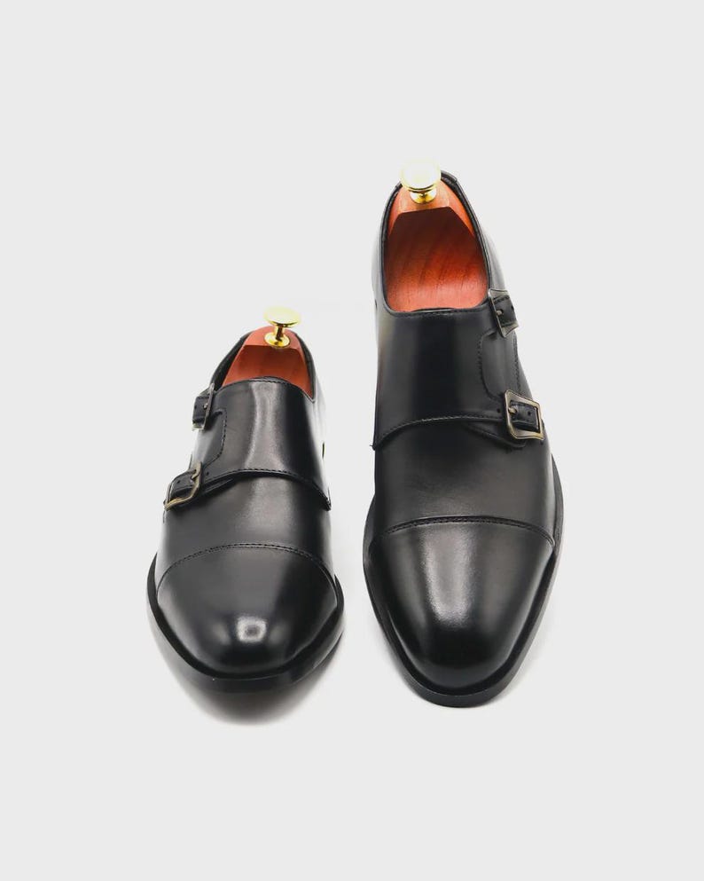 Puede incluir: Un par de zapatos negros de cuero con doble hebilla. Los zapatos tienen un dise&ntilde;o de puntera y dos hebillas ajustables. Hormas de madera en el interior. Se muestran sobre un fondo blanco. Estilo cl&aacute;sico.
