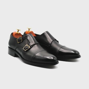 Puede incluir: Un par de zapatos negros de cuero con doble hebilla. Los zapatos tienen un dise&ntilde;o de puntera y hebillas doradas. Se insertan hormas dentro de los zapatos. Los zapatos est&aacute;n sobre un fondo blanco.