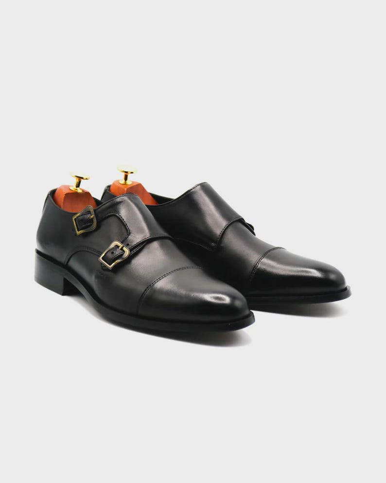 Puede incluir: Un par de zapatos negros de cuero con doble hebilla. Los zapatos tienen un dise&ntilde;o de puntera y hebillas doradas. Se insertan hormas dentro de los zapatos. Los zapatos est&aacute;n sobre un fondo blanco.