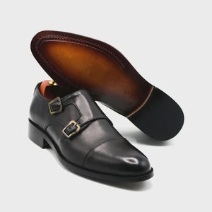 Puede incluir: Zapatos negros de cuero con doble hebilla y hebillas doradas. La suela es marr&oacute;n con un borde cosido. Un zapato est&aacute; en posici&oacute;n vertical, mientras que el otro est&aacute; en &aacute;ngulo para mostrar la suela.