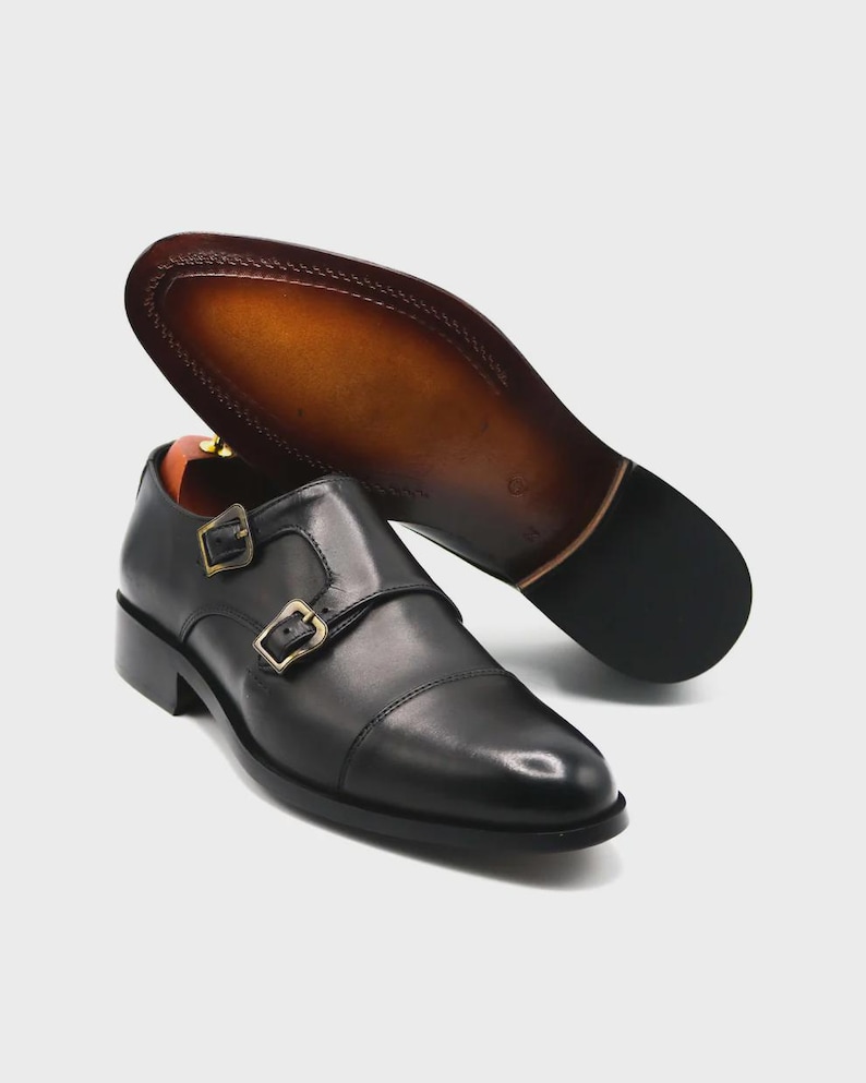 Puede incluir: Zapatos negros de cuero con doble hebilla y hebillas doradas. La suela es marr&oacute;n con un borde cosido. Un zapato est&aacute; en posici&oacute;n vertical, mientras que el otro est&aacute; en &aacute;ngulo para mostrar la suela.