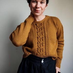 Peut inclure: Pull en tricot jaune moutarde avec un motif en tricot torsadé au centre. Le pull a des manches longues et un ourlet côtelé. La personne porte une jupe noire. Le pull est un vêtement chaud et confortable.